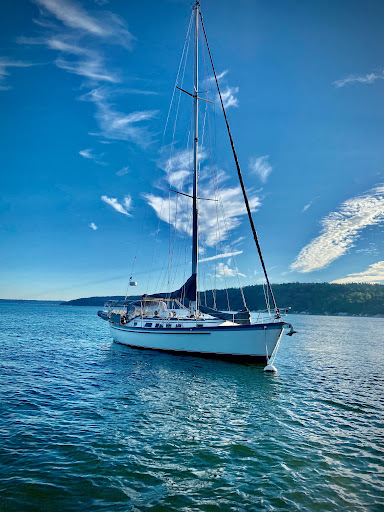 A large sailboat in blue water, under a deep blue sky with wispy white clouds near the horizon. A stretch of land, covered in green trees, is visible in the background, on the horizon.