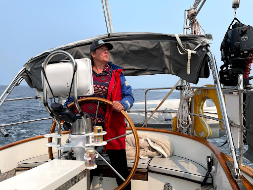 A woman standing at the helm of a sailboat, with her hands on the boat's wheel. She is facing the camera, and looking up and to her left. She has light skin and is wearing a baseball cap, a blue-and-red-patterned sweater, and a blue-and-red-patterned raincoat.
