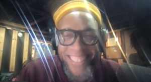 Speech in a zoom window. He has on black glasses and headphones over his ears. He's wearing a yellow beanie.