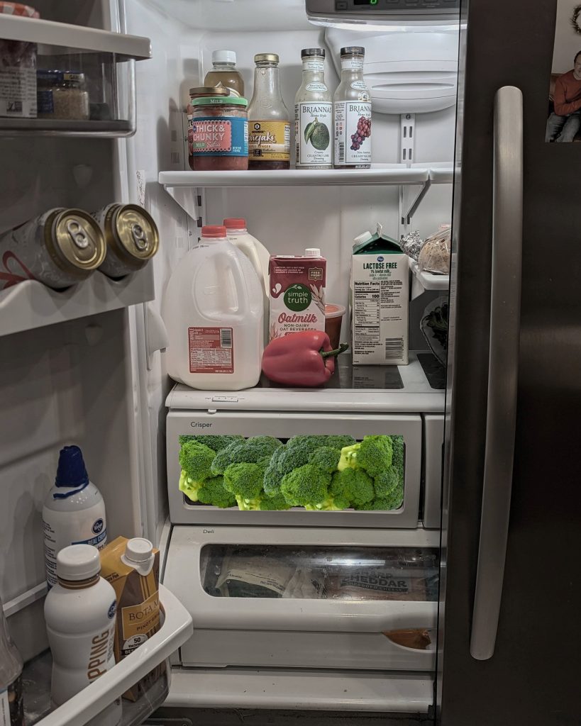 A vertical photo of a refrigerator with its left door open. There are jars of sauce and dressing on the top shelf, cartons of milk and a red bell pepper on the bottom shelf, and cans of beer and other beverages on the shelves in the door. The crisper drawer, below the bottom shelf, has been Photoshopped to appear as though it's full to bursting with green heads of broccoli.