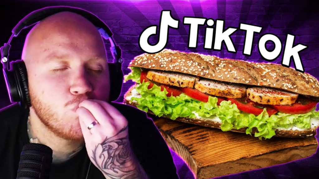 A thumbnail image from a YouTube video. On the left of the frame, there is a bald white man with a red moustache and beard. He is wearing headphones, his eyes are closed, and he is kissing his pinched fingers on his left hand. To the right of the frame, there is a cutout image of a sub sandwich with lettuce, tomato, mayonnaise and roasted chicken on multigrain bread. Above the sandwich, the TikTok logo is visible. The background is a brick wall bathed in violet light.