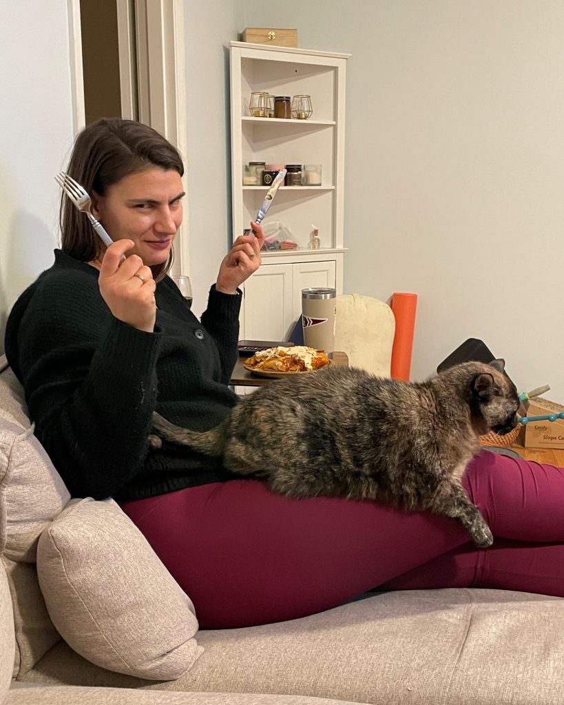 A woman sitting on a couch with her legs out in front of her. She is holding a butter knife in her left hand and a fork in her right hand, and she is holding up her hands in mock resignation. There is a tortie cat in her lap.