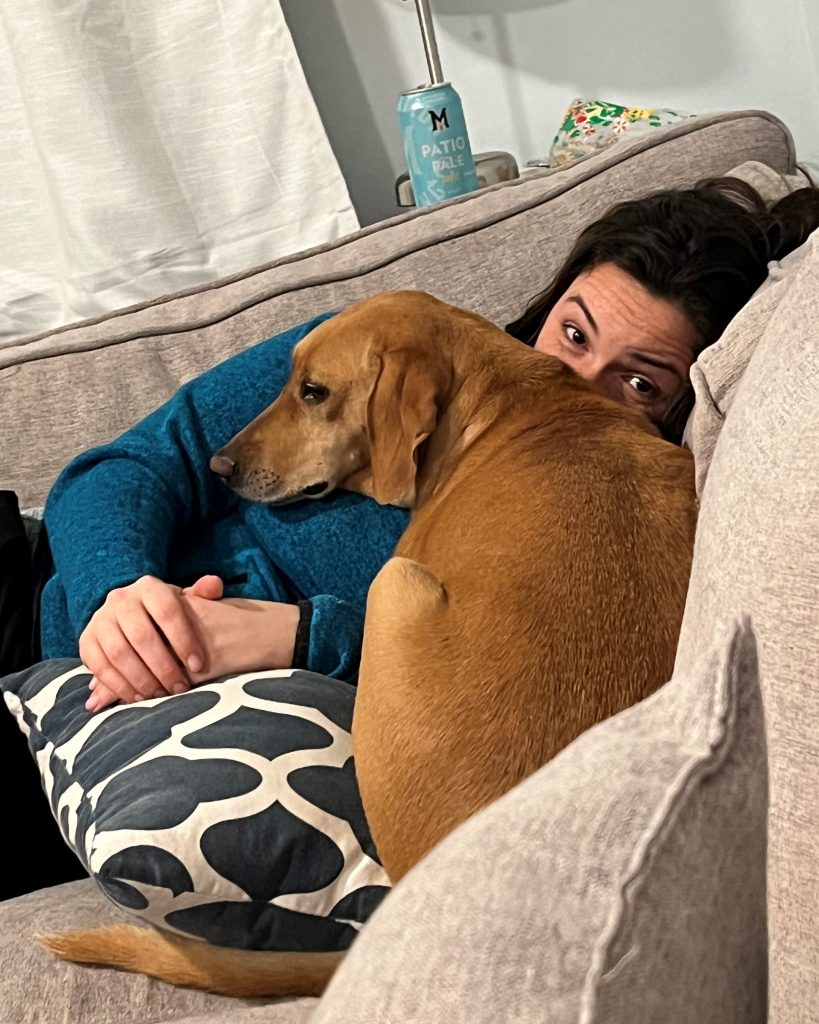 A woman lying on a couch with a big brown hound-type dog snuggled up into her chest. The woman is looking at the camera and the dog is staring off into space.