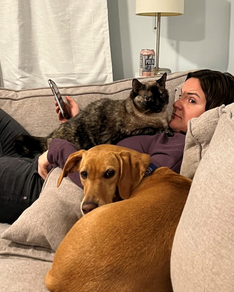 A woman lying on her back on a couch, with a dark-colored tortie cat lying on her chest. A brown dog is curled up beside them. All three are looking at the camera.