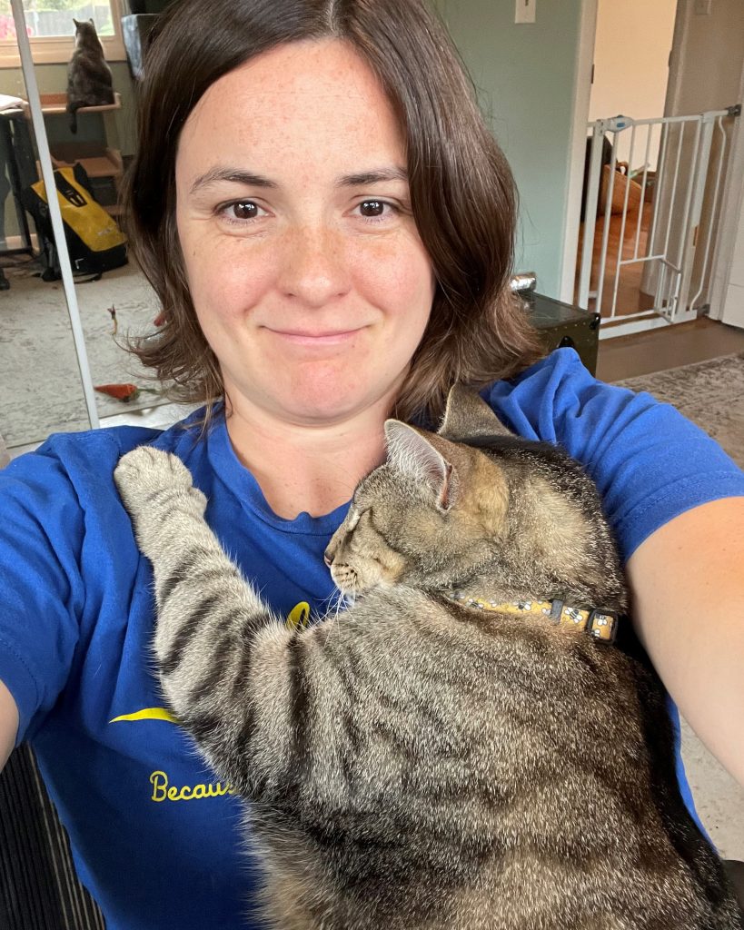 A woman taking a selfie with a grey tabby cat. The cat is snuggled into her chest with his eyes closed, and has one paw resting on the woman's shoulder.