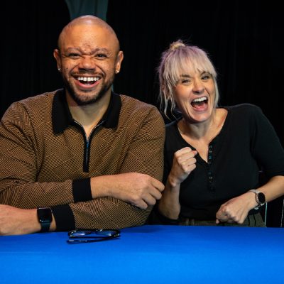 Jarrett Hill and Brea Grant. Jarrett is wearing a brown collared sweat shirt and Brea is wearing a black top. They both are sitting a table with a blue tablecloth and have on big smiles.