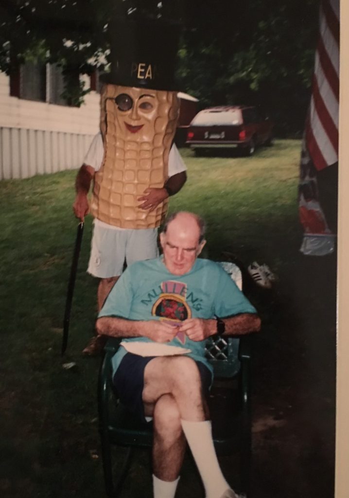 An old film photograph. An old, balding man is sitting on a lawn chair under a tree. His legs are crossed, he has a plate on his lap, and he is wearing a blue shirt that says 