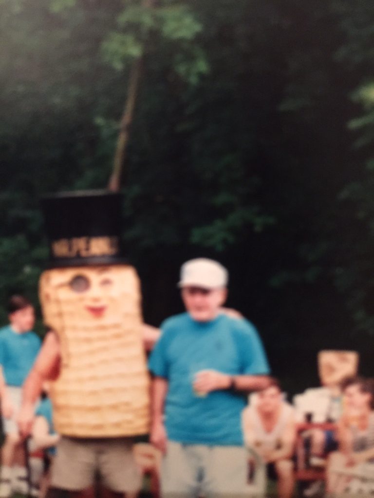 An old, out-of-focus film photograph. Two men are standing together. The man on the left is wearing a costume of Mr. Peanut, which covers his torso. The costume is a peanut with a monocle, bright red lips, a cane, and a top hat that reads 