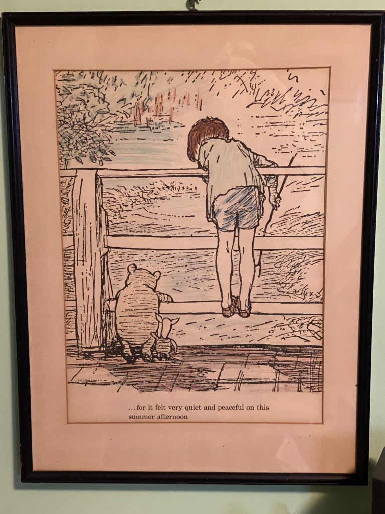 A close-up photo of a framed print, hanging on a wall. The print is a scene from one of the Winnie-the-Pooh books and shows Pooh, Piglet and Christopher Robin. They are standing side by side on a bridge, facing away from the viewer, and dropping sticks into a river. Below, the text reads: 