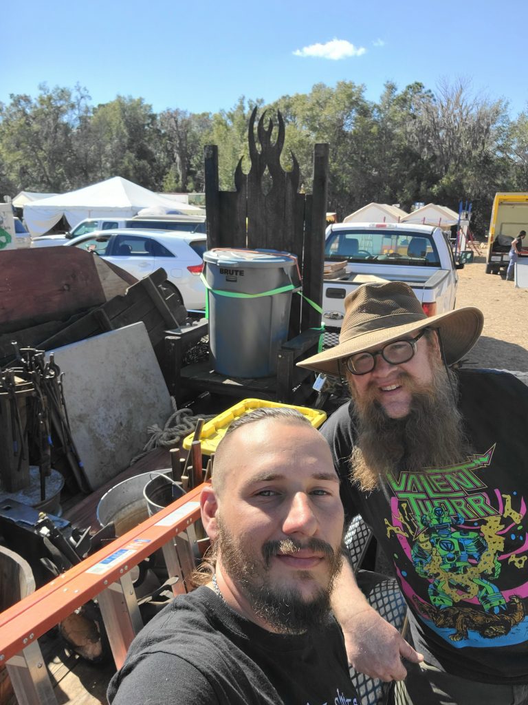 A selfie of two men outdoors in daylight in front of a trailer, loaded with blacksmithing materials and products
