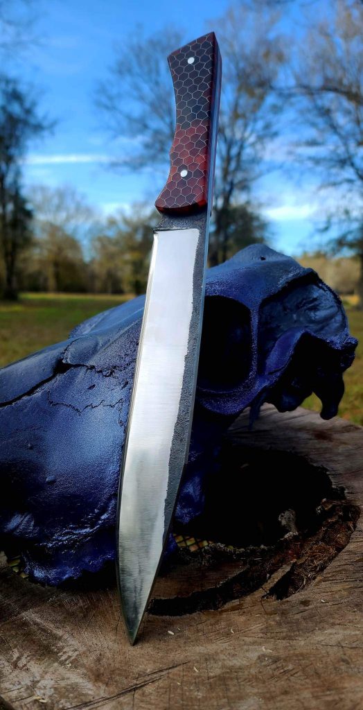 A handmade knife with a long, narrow blade. It is displayed on a tree stump, leaning point-down against a blackened, burned cow skull. The hilt is red and black, and has a texture similar to a snake's scales.