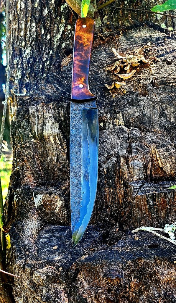 A handmade knife, laid flat against a tree trunk. The blade appears to be about four or five inches long.