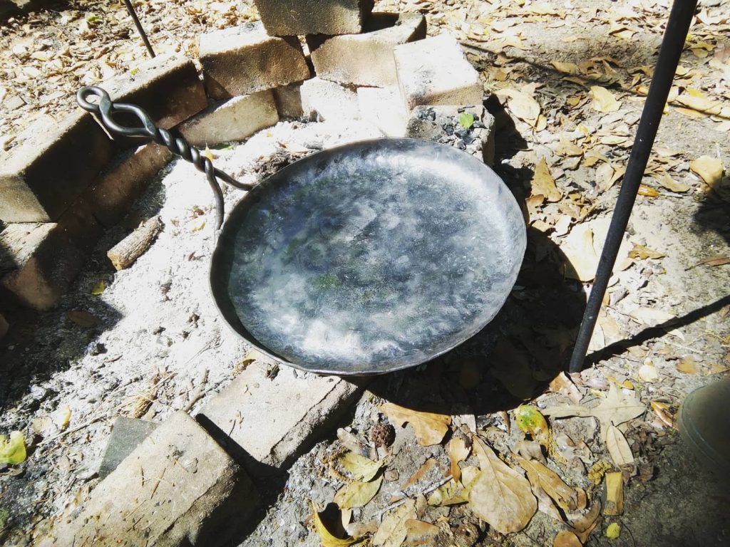 A handmade cast-iron skillet, displayed on what appears to be a brick fire pit. The pan is wide and shallow. The fire pit is surrounded by yellowed autumn leaves.