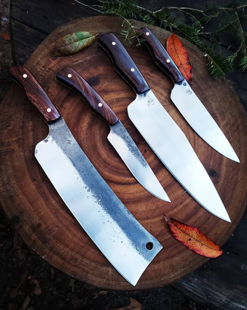 Four matching handmade kitchen knives, displayed next to each other on a tree stump. There is a butcher knife, a paring knife, a full-sized chef's knife, and a smaller chef's knife. The hilts are made of a beautiful dark wood. On each blade, near the hilt, there is a set of Anglo-Saxon runes that translate to 
