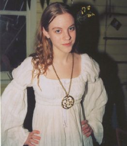 Maddy Myers as a teen, wearing a lovely white dress with poofy sleeves. She has long hair and a large statement necklace