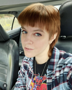 Maddy Myers as an adult. She's wearing a colorful flannel with a t-shirt beneath. Her hair is MUCH shorter than her teen years. 