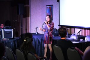 Helen Hong performing stand-up comedy! She's wearing a dress with blue, red and white vertical stripes. There a few people in the very front row looking at her. She has a microphone in her hand. 