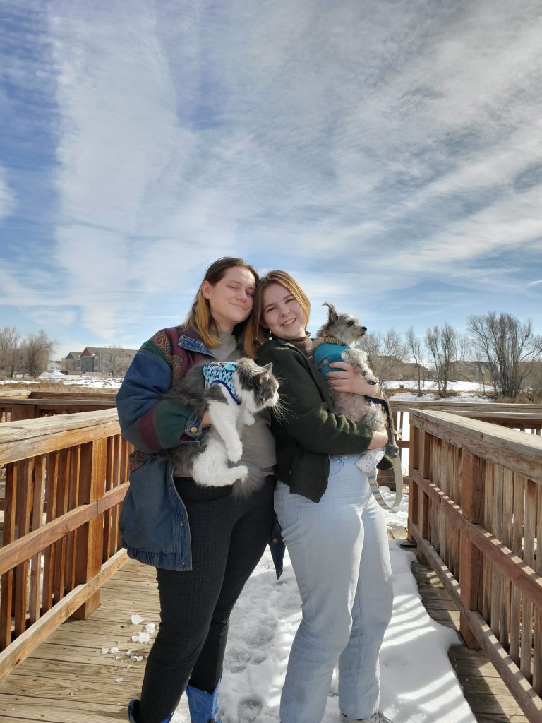 Two young women stand together on a snow-covered wooden bridge. The woman on the left has shoulder-length brown hair and is holding a gray-and-white cat in her right hand. The cat is wearing a blue patterned vest. The woman on the right is wearing a black jacket and is holding a scruffy gray three-legged terrier.