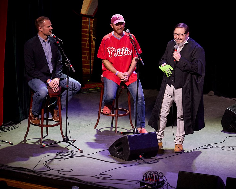 Three men on a stage, speaking into microphones. The man on the left has close-cropped hair and is wearing a dark blazer and jeans. He is sitting on a stool. The man in the middle is wearing a red Phillies baseball cap, a red Phillies jersey, blue jeans, and red Crocs. He is sitting on a stool. The man on the right is John Hodgman, wearing a black judge's robe and gray jeans. He is standing and holding a pair of green gloves in his right hand.
