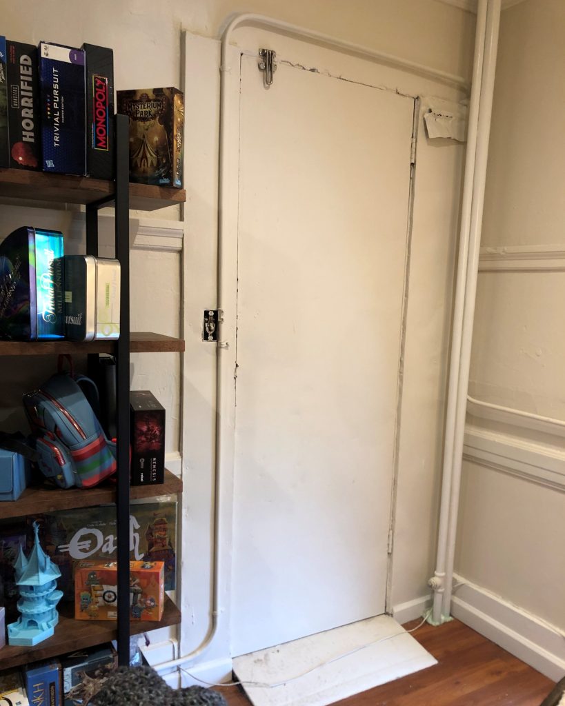 A closed white door, with no handle or doorknob on the visible side of it. A hotel-style folding bolt is holding it shut from the top left corner. To the left of the door, there is a bookshelf laden with board games such as Monopoly, Horrified, and three different editions of Trivial Pursuit.