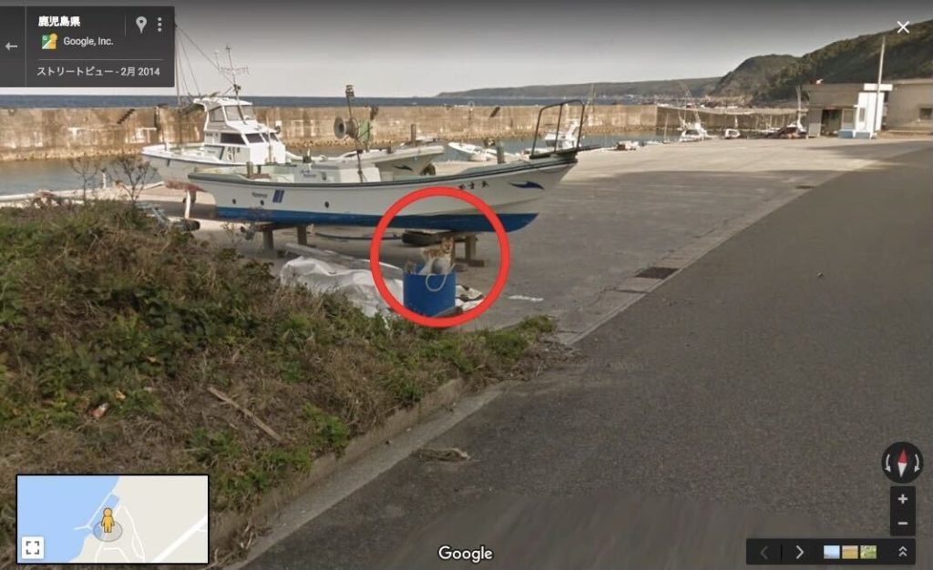 A screenshot of Street View on Google Maps, showing a view of a concrete dock in Japan. There is a river in the background, with boats sailing up and down it. There is a white boat with blue trim up on blocks, and an excited Shiba Inu dog standing under it. There is a bright red circle around the dog.