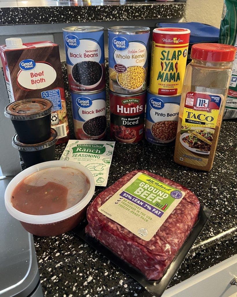 An array of ingredients displayed on a black countertop. The ingredients are beef broth, two cans of black beans, one can of corn, one can of diced tomatoes, one container of 