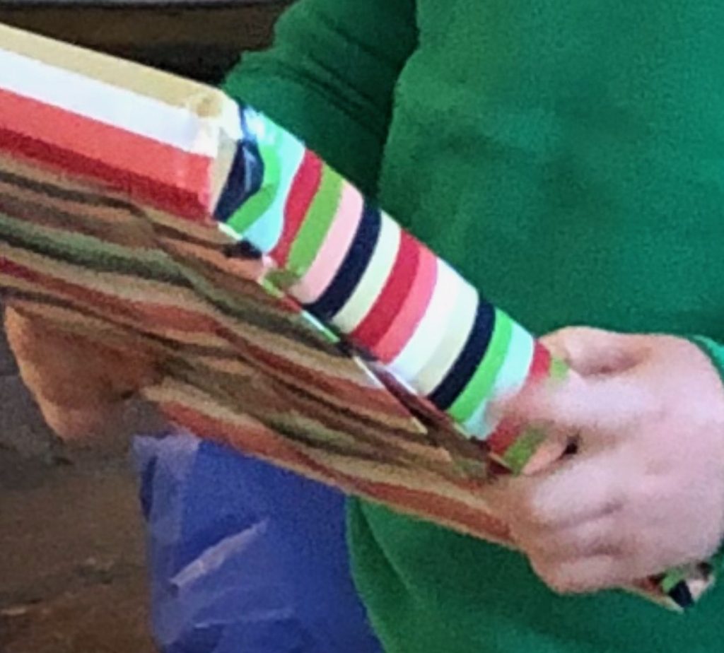 A close-up of a person in a green sweater holding a rectangular wrapped gift. The wrapping paper is patterned with stripes of green, light blue, red, dark green, pink, black, white, red, and two shades of off-white.