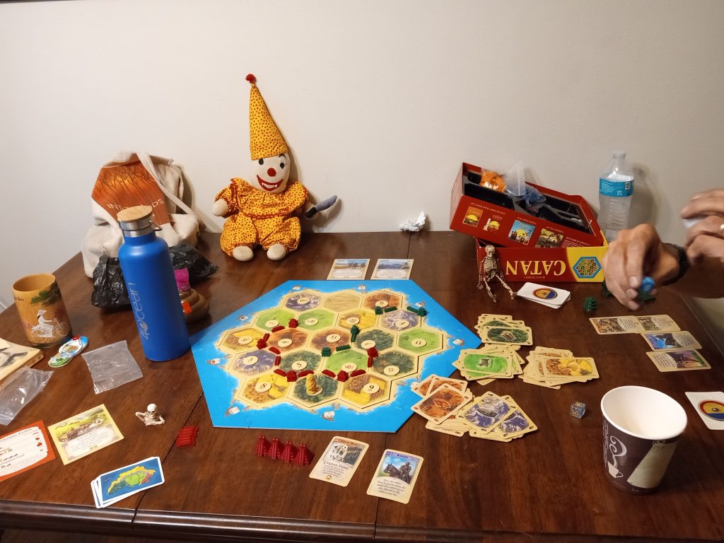 The board game Settlers of Catan set on a table, with the stuffed clown propped up next to it, some toy skeletons on the table, the old coffee cup and other water bottles