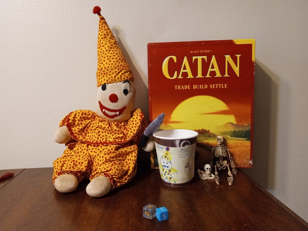 The stuffed clown, the coffee cup with its image on it, two dice, and the game Settlers of Catan propped up behind the cup