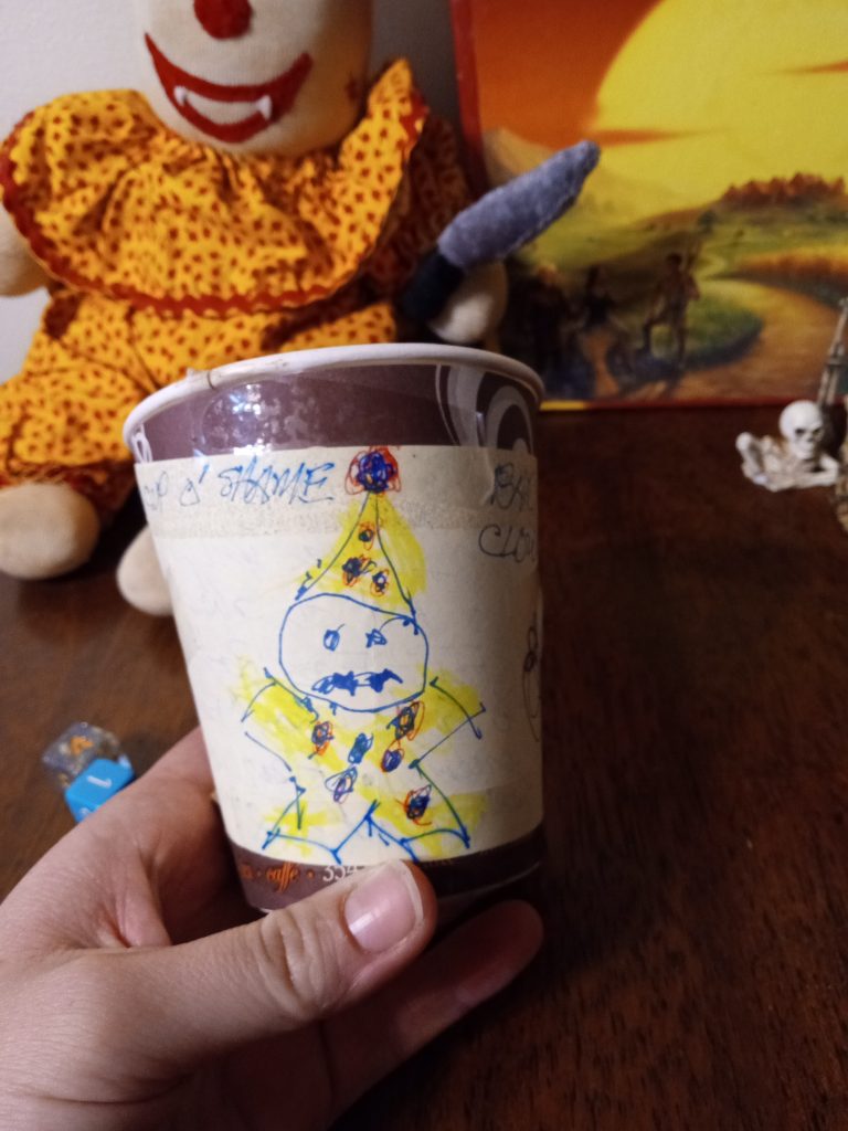 a hand holding a paper coffee cup with a drawing of a creepy clown on it