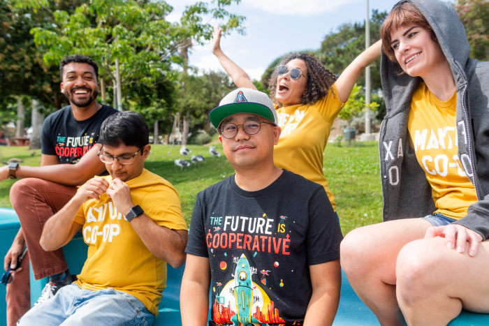 Five MaxFun worker-owners posed across a bench in a park wearing MaxFun co-op themed merch.