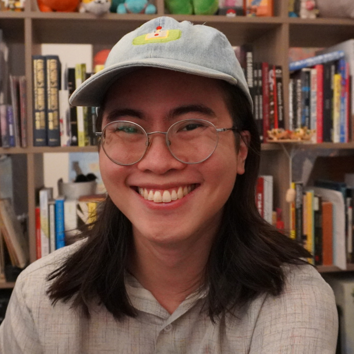 Tom Lum smiling at the camera wearing a hat over his long hair and sitting in front of a bookshelf