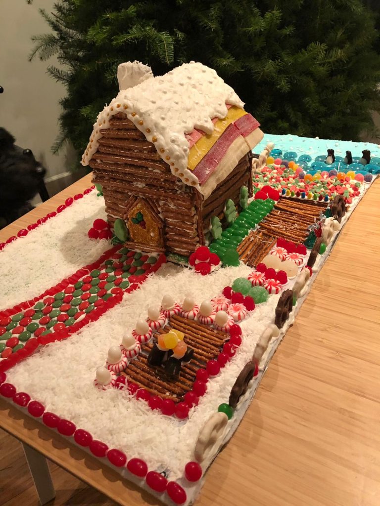 A gingerbread house with colorful candy, fire pit in front, and penguins in the back on a blue icing lake