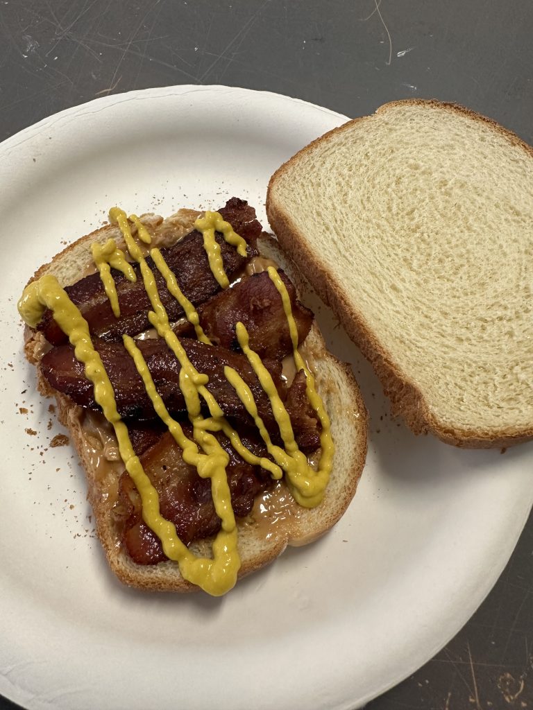 A sandwich opened with a layer of peanut butter on the bottom, then bacon, with yellow mustard drizzled on top