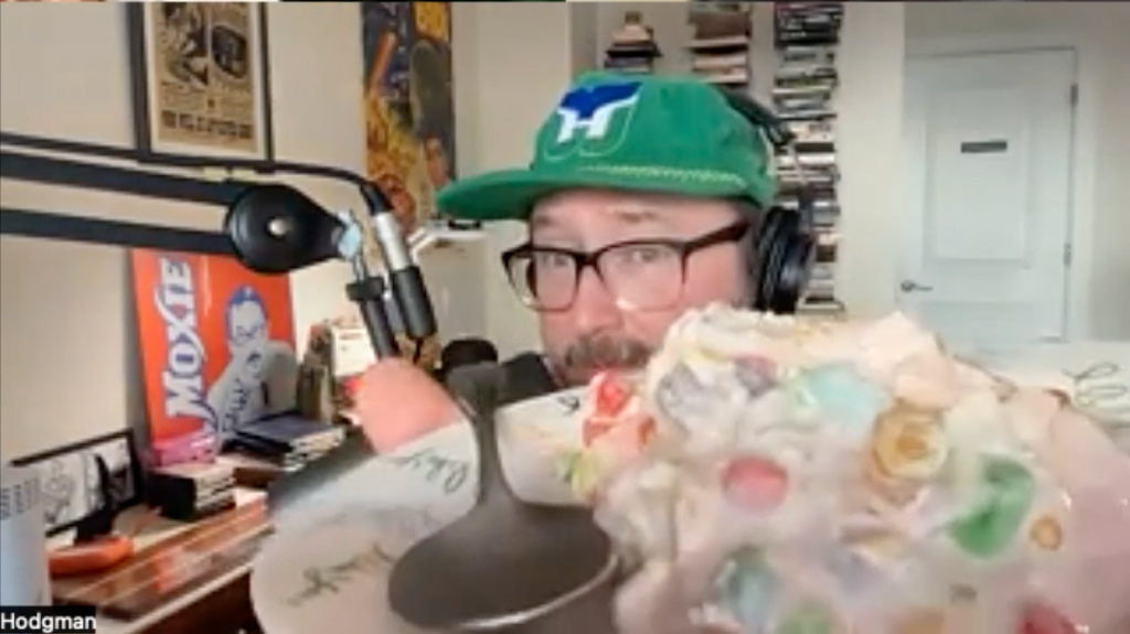 A video conference screenshot of a man holding a plate of a creamy looking food with multi color mini marshmallows in it