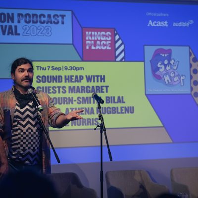 John-Luke Roberts (a white man with brown hair and a moustache) stands onstage in front of a slide with the name of the podcast and its guests on it, in front of a microphone.