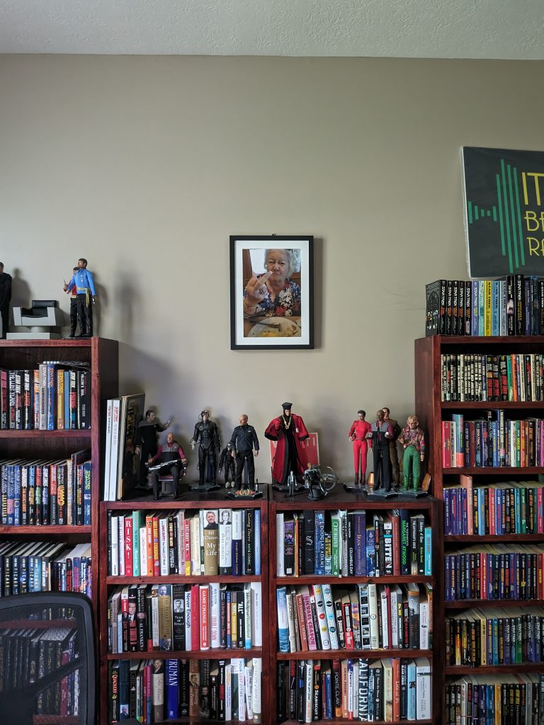 A large framed photo of an older woman with her middle finger up, hanging on a wall surrounded by full bookshelves