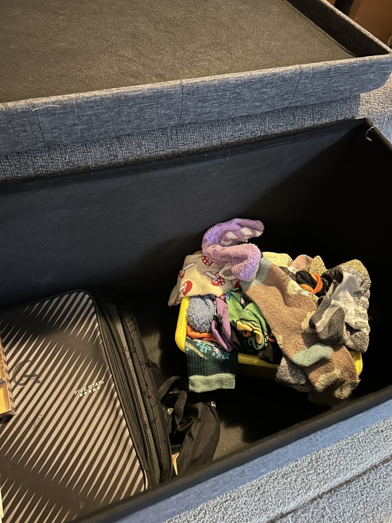 A storage ottoman open with a yellow bin of socks inside, next to a school binder