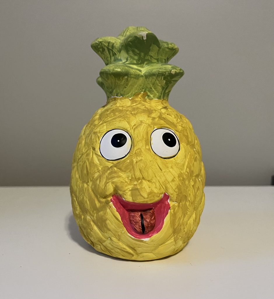 A painted ceramic cartoonish-looking pineapple piggie bank, with eyes and a smiling mouth