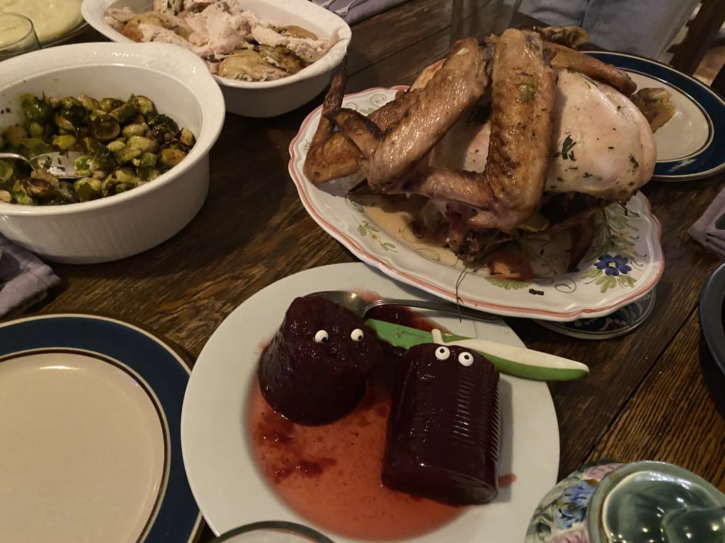 Two canned cranberry sauces out of cans on a plate, with candy eyes on them. The plate with the sauces is on a table with other Thanksgiving foods