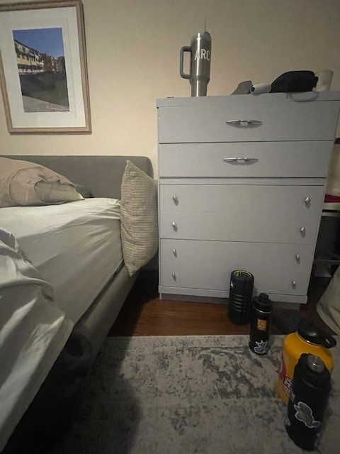 A bed with a tall dresser to the right of it. A decorative pillow is propped up in the space between bed and dresser. Three water bottles are in the right foreground.