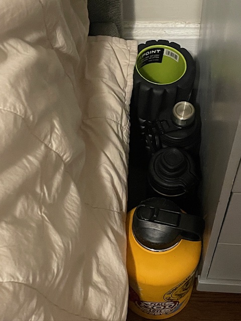 Three reusable water bottles and a foam roller standing on the floor between a bed and dresser