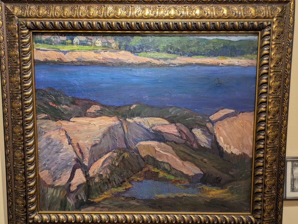 An oil landscape painting showing rocks in the foreground and a lake in the background