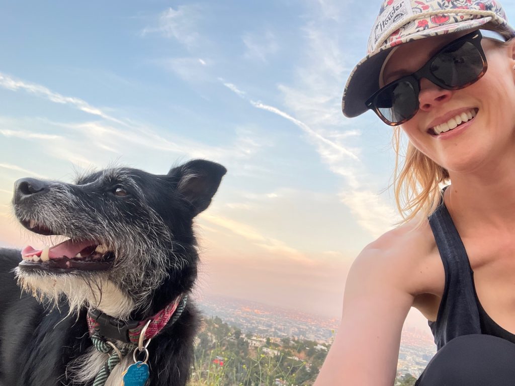 A black scruffy dog with some white fur on the snoot and chest sitting next to guest Janet Varney on a hike