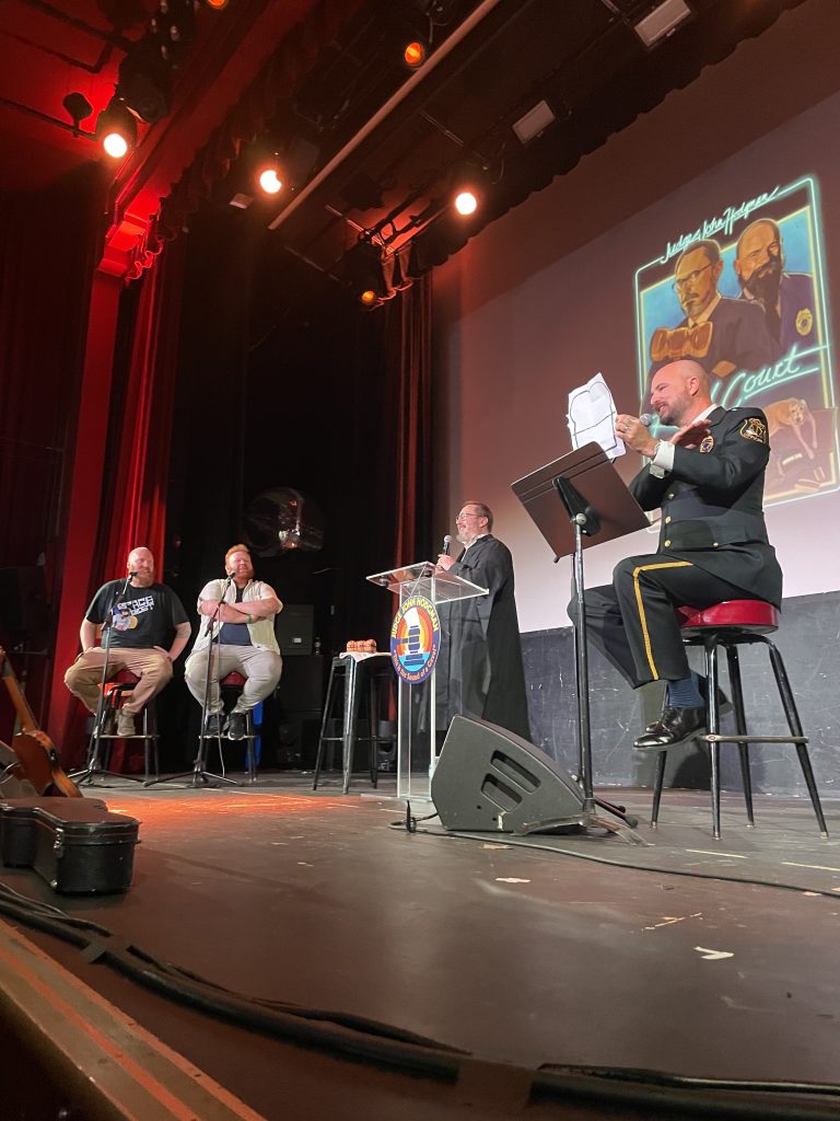 Four people on a stage. One person is holding a white piece of paper with a drawing in black sharpie of a sandwich with a horizontal line through the middle