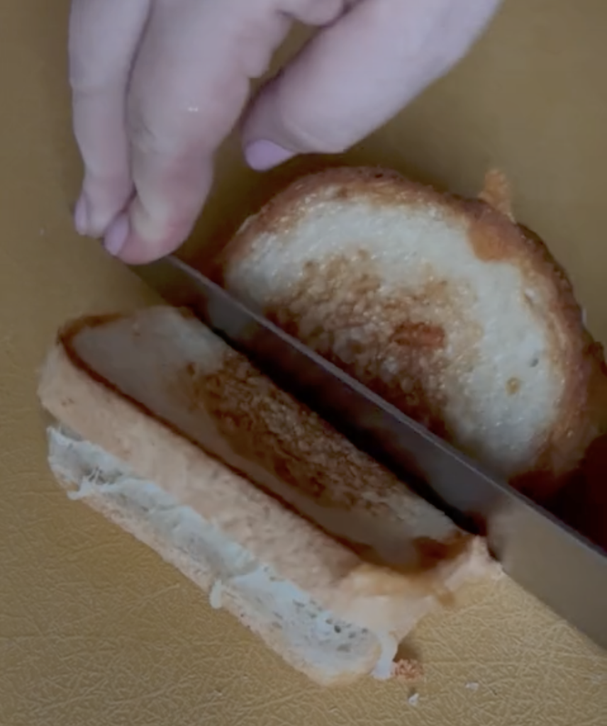 A grilled cheese sandwich on white bread on a wooden cutting board, with a person's hands using a knife to cut through the middle horizontally