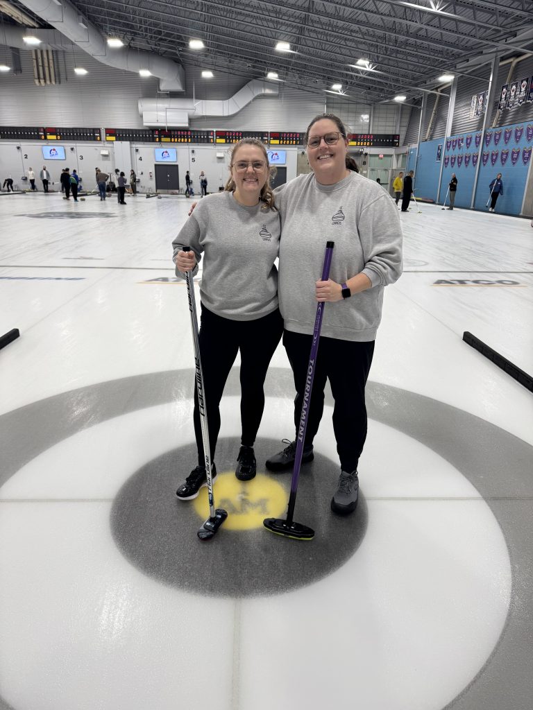 Two people standing on the ice of a curling rink, both holding curling brooms