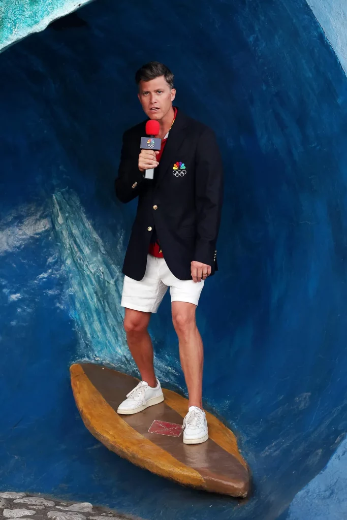 A man wearing a blazer and white shorts standing on a surfboard in a fake wave