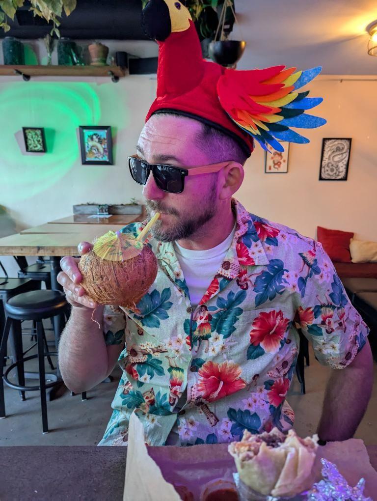 A person drinking a drink while wearing a parrot hat and wearing a tropical themed shirt