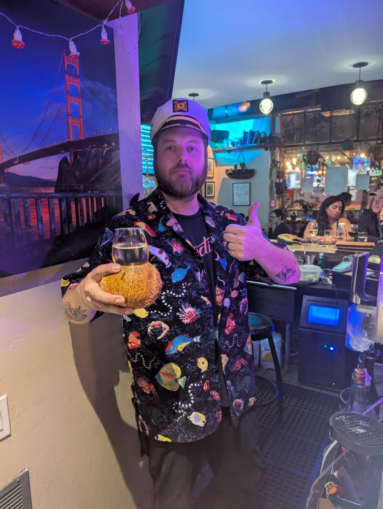 A person wearing a captain's hat and tropical themed shirt, holding a drink in one hand and giving a thumbs up in the other hand